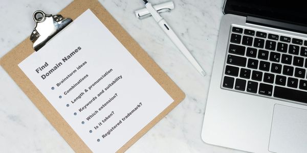 Clipboard with domain name brainstorming tips beside a laptop and pen.