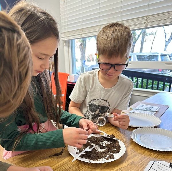 Children exploring soil with magnifying glasses and spoons indoors.