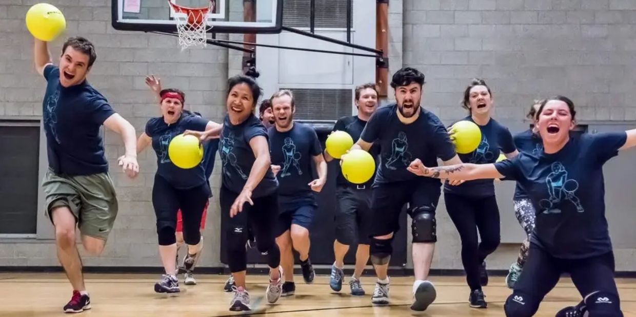Group of people playing dodgeball indoors with yellow balls, running energetically towards the camera.