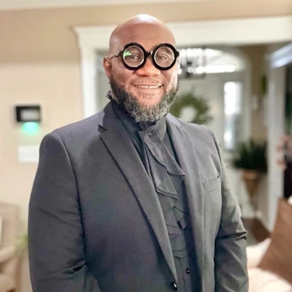 Man in a black suit with round glasses smiling indoors.