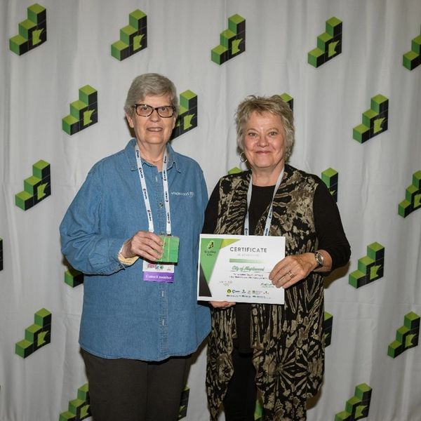 Two women holding a certificate and an award at an event with a branded backdrop.