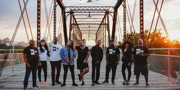 Group of nine people posing on a bridge at sunset, wearing matching "ONE" shirts.