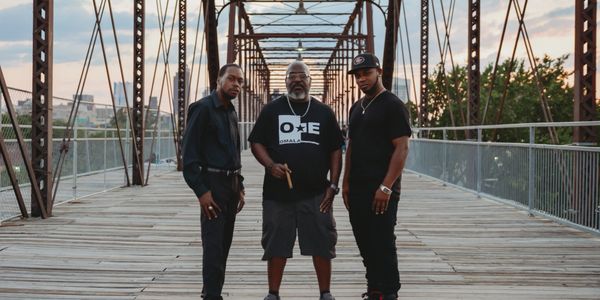 Three men stand confidently on a wooden bridge at sunset.