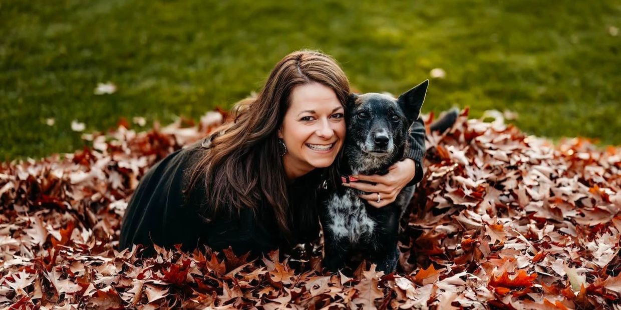 Woman joyfully hugging her dog amidst autumn leaves in a park.