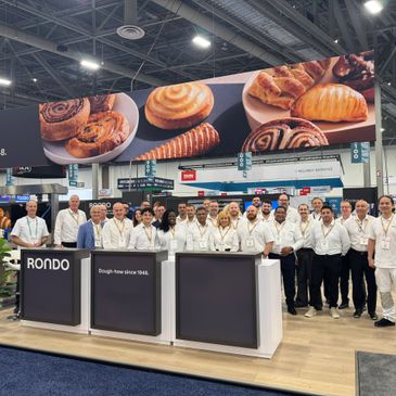 Group of people in white uniforms posing at a bakery trade show booth.