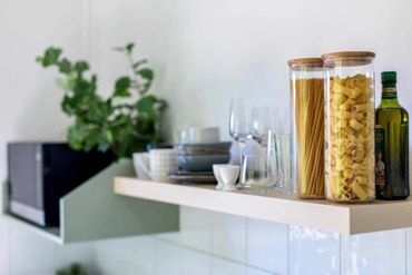 Kitchen shelf with pasta jars, olive oil, glasses, and bowls.