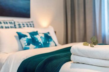 Cozy bedroom with white linens, teal pillows, and neatly folded towels with a small leafy decoration.