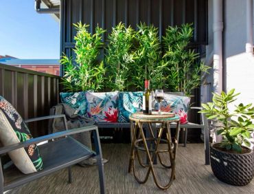 Cozy balcony with tropical cushions, greenery, and a wine setup on a wicker table.