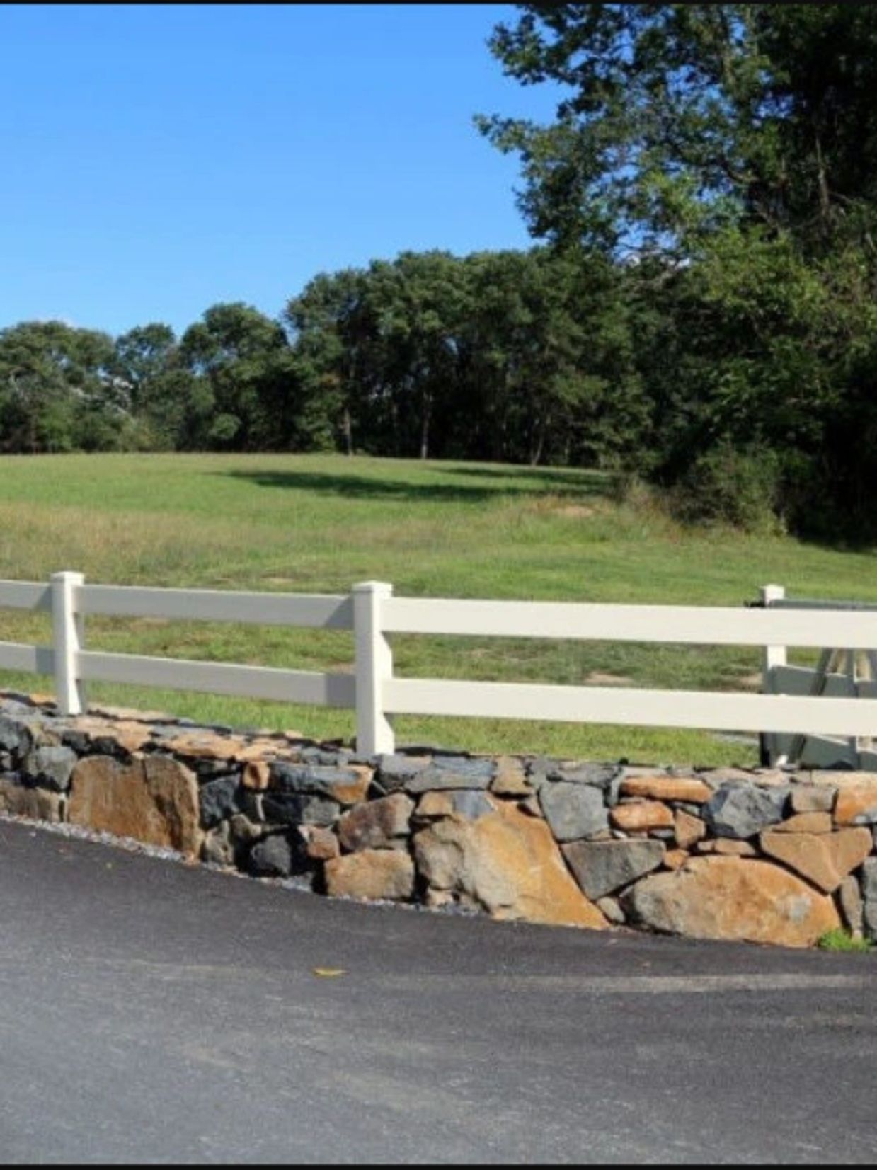 A white fence and stone wall border a grassy field with trees under a clear blue sky.