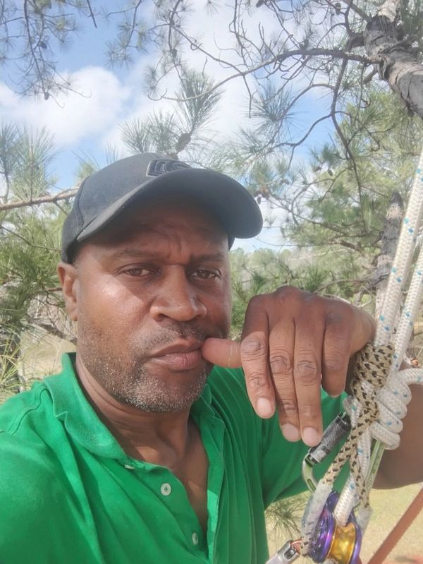 Man wearing a green shirt and black cap outdoors among trees with climbing gear.