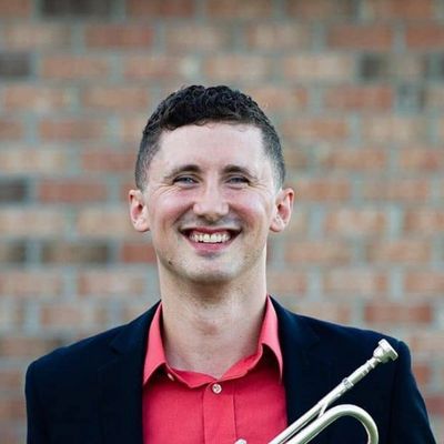 Smiling man holding a trumpet, dressed in a red shirt and dark blazer.