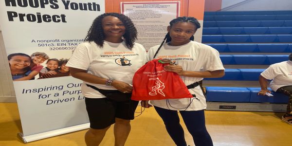 Two women in HOOPS Youth Project shirts posing with a red bag in a gymnasium.