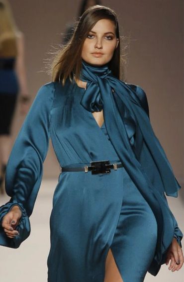 Model wearing a sleek teal satin dress with a matching scarf and black belt on runway.