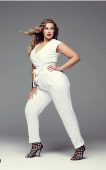 Confident woman in white jumpsuit and black heels poses against gray background.