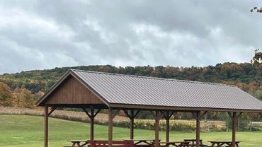 A wooden pavilion with picnic tables in a green field under a cloudy sky.