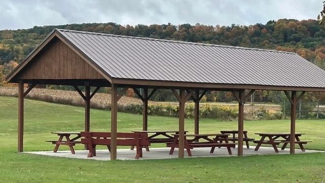 A wooden pavilion with picnic tables in a green field under a cloudy sky.