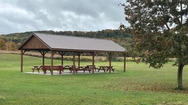A wooden pavilion with picnic tables in a green field under a cloudy sky.