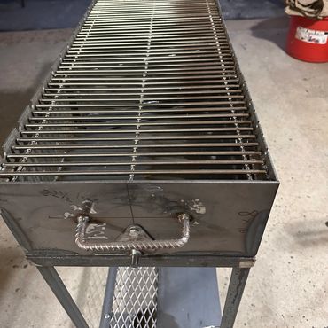 Metal grill with sturdy handle and legs, designed for outdoor cooking.