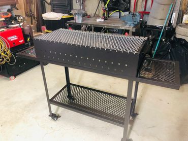 Custom metal charcoal grill with side shelves on wheels in a workshop.