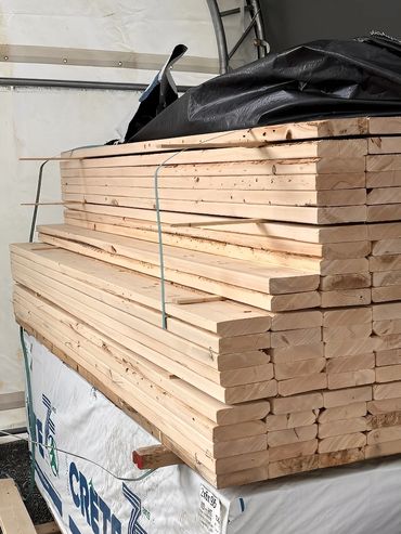 Stacked lumber planks with spacers and black tarp, stored inside a shelter.