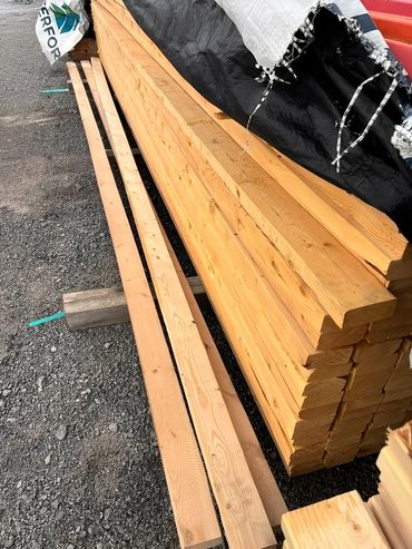 Stacked wooden planks covered with tarp, resting on gravel ground outdoors.