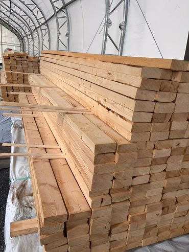 Stacked lumber planks with spacers, stored inside a metal-framed shelter.