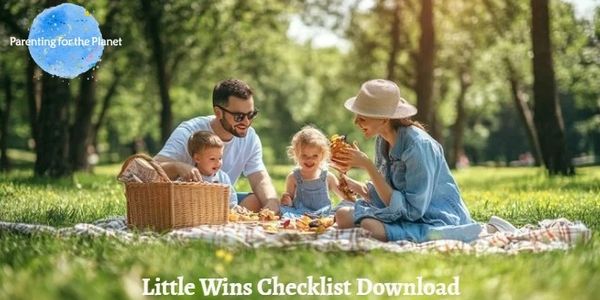 A mother and father sit with two young children on a picnic rug with a basket under tall green trees