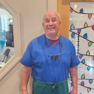 Smiling man in blue scrubs stands indoors near decorated glass door.