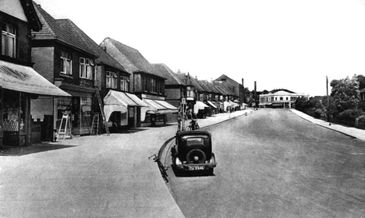 Black and white photo of an old street with vintage cars and small shops.