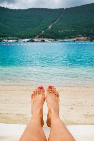 Relaxing feet with pink nail polish by the beach and clear blue water.