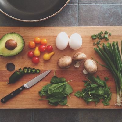 Chopping board on counter with avocado, eggs, spring onion, mushrooms, tomatoes. Frying pan behind