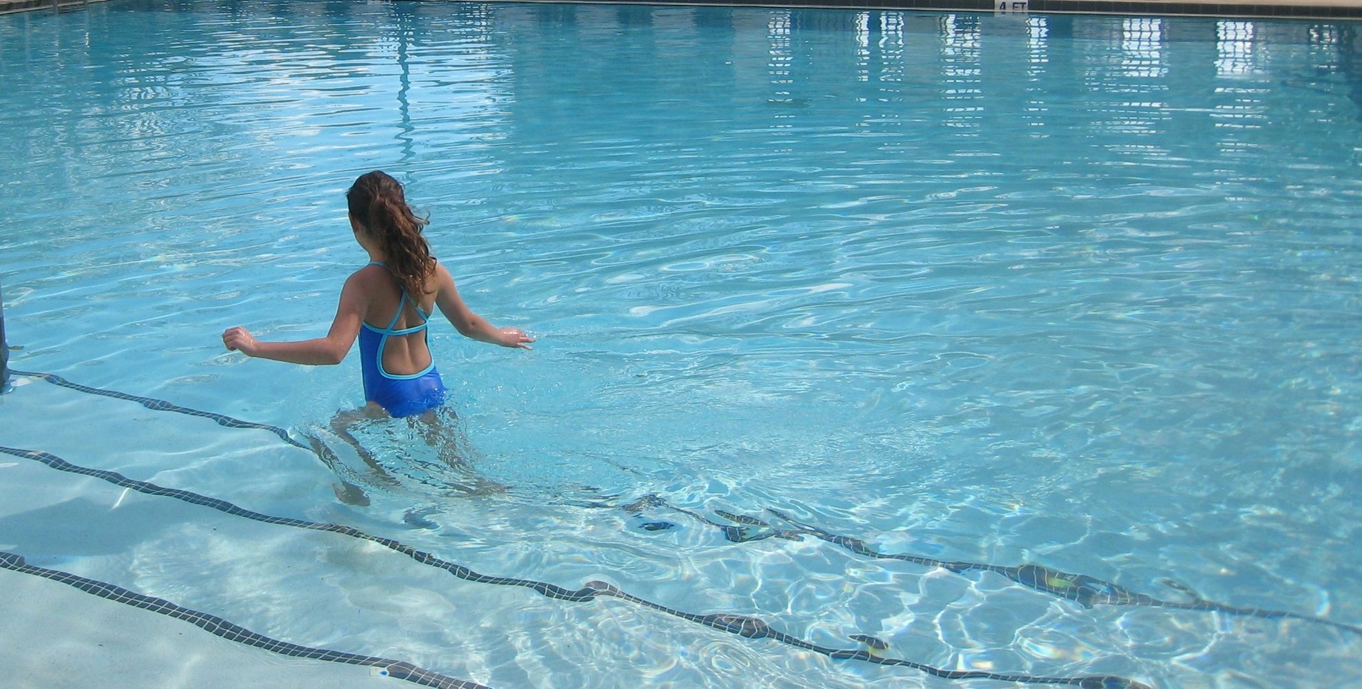 Girl wading into a pool wearing a blue and aqua swimsuit.