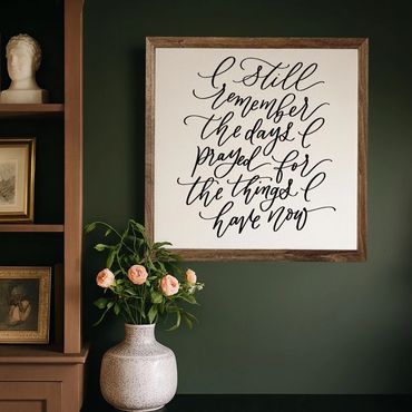 Framed calligraphy quote with flowers in a ceramic vase against a dark green wall.