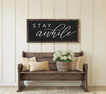 Rustic bench with cushions and flowers beneath a 'Stay Awhile' sign.