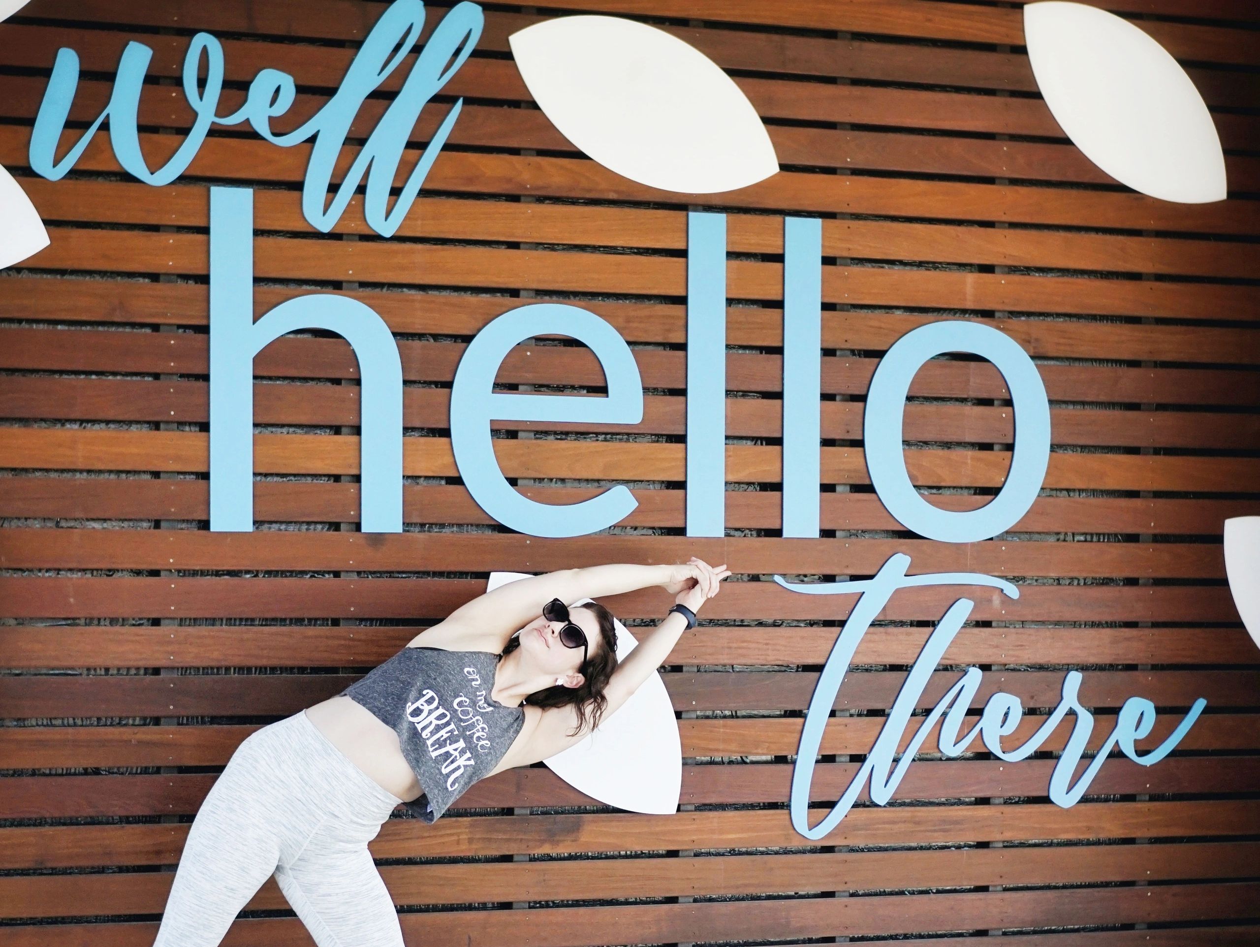 Yoga teacher in triangle pose pointing to a large HELLO sign on a wooden wall outdoors