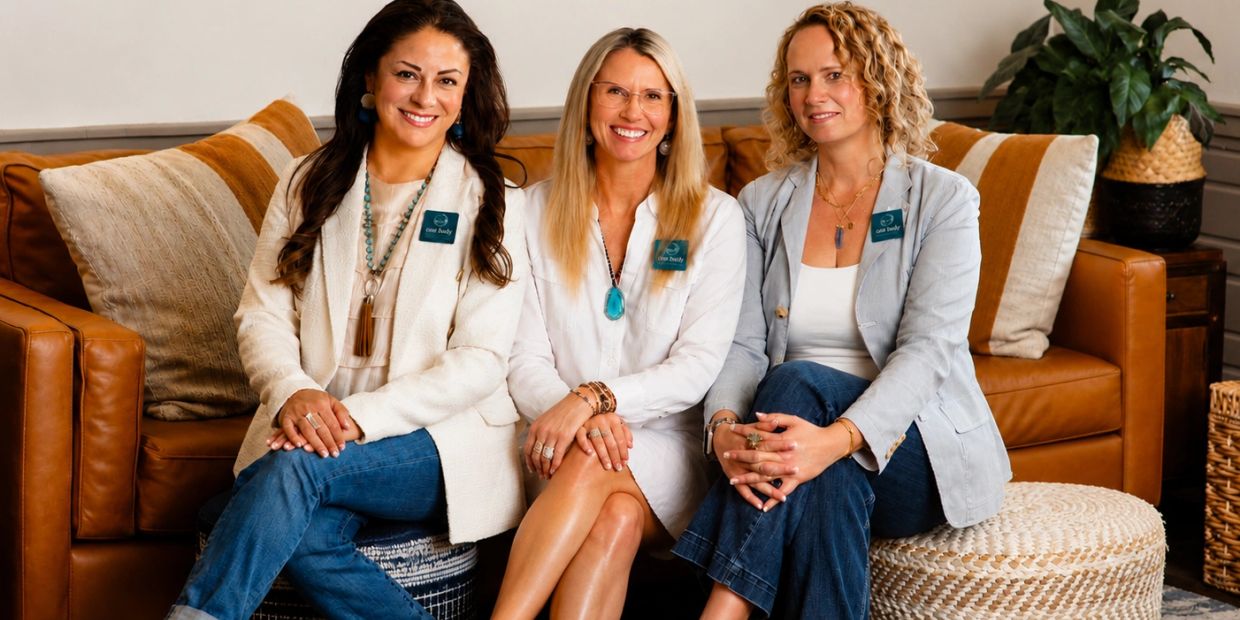 Three women smiling while seated on a brown leather couch in a cozy living room.