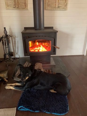 A black dog lying on a blue mat by a glowing wood stove.