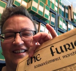 Jackie Rhodes smiles broadly while holding a copy of THE FURIES newspaper up to the camera.