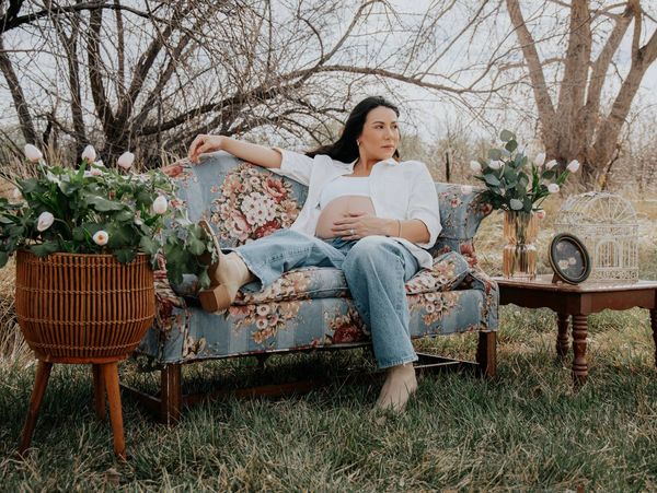 Pregnant woman relaxing on a floral couch outdoors with plants and vintage decor.