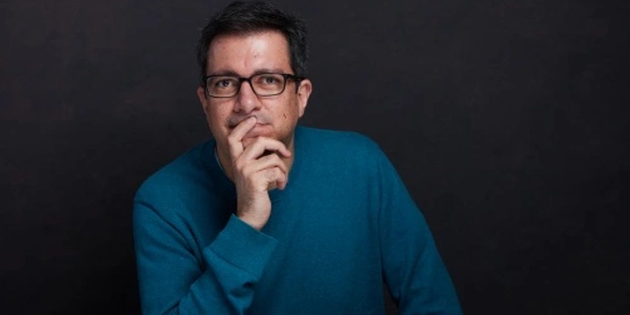 Man in a blue sweater with glasses posing thoughtfully against a dark background.