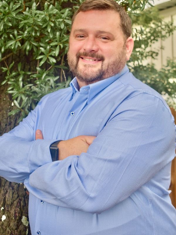 Smiling man in a blue shirt with crossed arms standing outdoors near a tree.