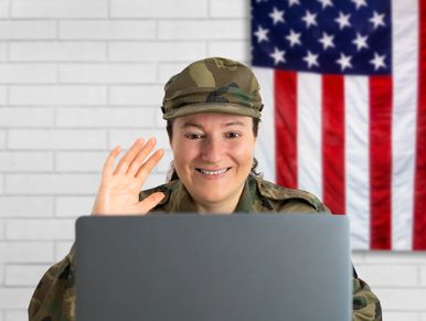 Smiling female soldier in uniform waving during a video call with an American flag in the background.