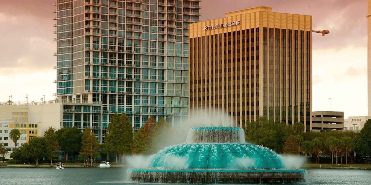 A large fountain in a city lake with skyscrapers and trees in the background.