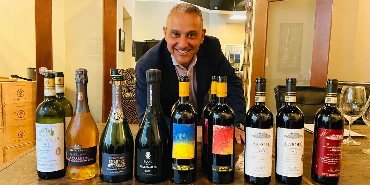 Man smiling behind a row of assorted wine bottles on a table.