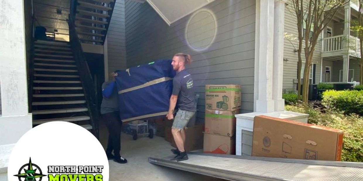 Two movers carrying a large wrapped item into a building with moving boxes nearby.