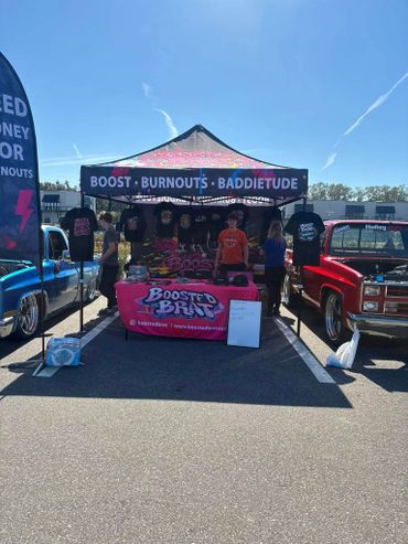 Car event booth selling Boosted Brat merchandise with classic trucks on each side.