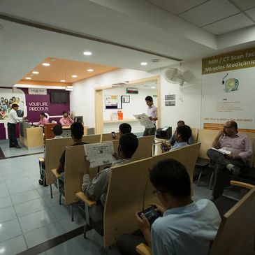 People waiting in a medical facility's seating area, some reading newspapers or using phones.