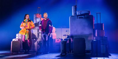 Two actors on stage with suitcases under blue light.