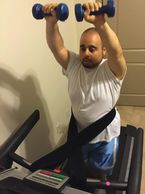 A man raising his arms while holding dumbbells while walking on a treadmill connecting himself with the treadmill safety waist. belt.