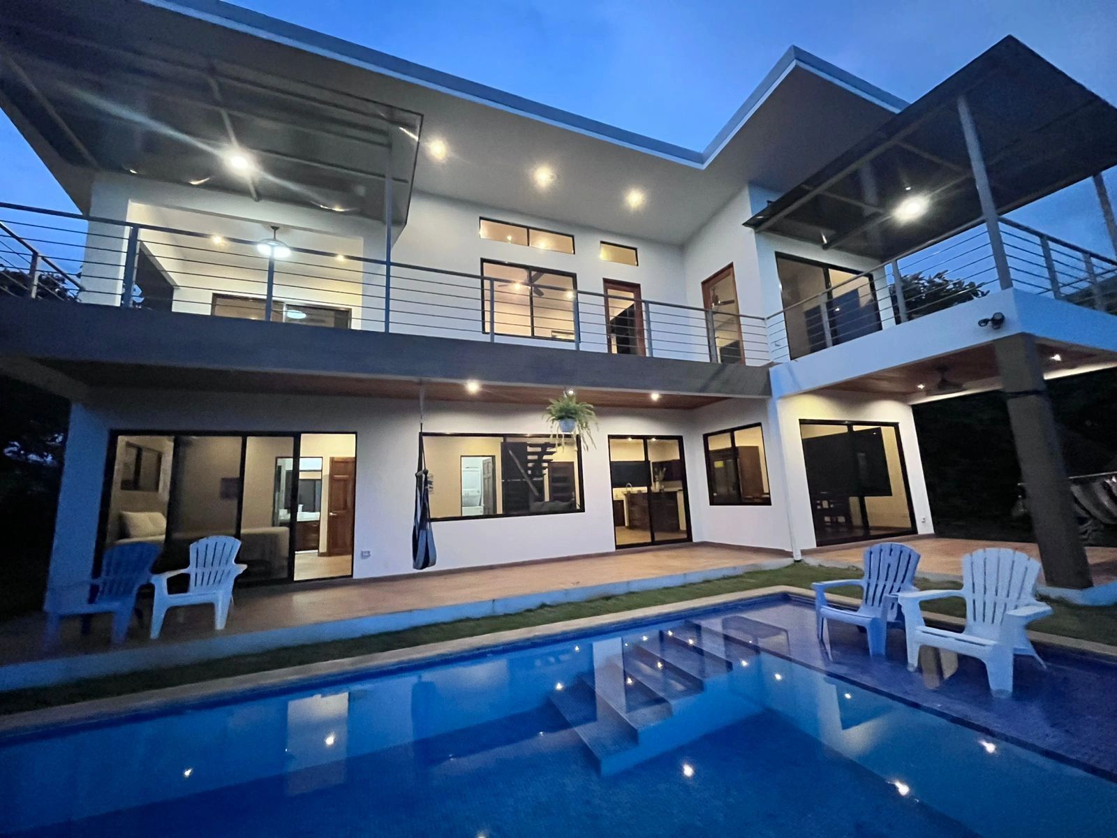 Modern two-story house with illuminated poolside patio at dusk.